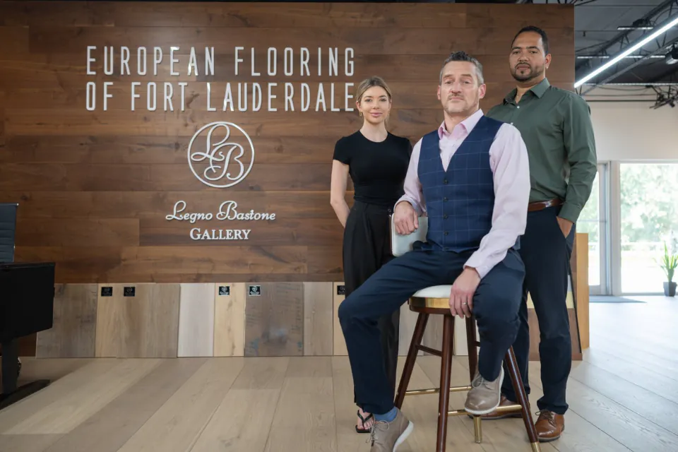 Three-person team posed in a flooring showroom with the company wall behind them